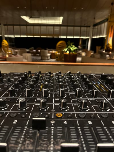 This is a close-up, angled shot of a black DJ mixer. The mixer is covered in various knobs, buttons, and sliders, with labels like "FILTER," "SEND," and "BEAT FX ASSIGN" visible. The faders have numerical markings from 2 to 5. The scene is dimly lit, with some warm light reflecting off the mixer and some yellow accents on the device itself. In the blurred background, there are hints of a dimly lit interior space with what appears to be furniture and possibly plants. The overall impression is that of a professional audio setup in a relaxed, possibly evening setting. There are no people visible, and the focus is entirely on the intricate details of the DJ equipment.