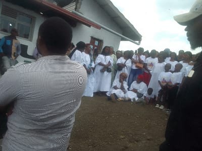 A large group of people, mostly wearing white clothing, gather outdoors in front of a building. Some individuals are sitting on the ground. Several children are wearing t-shirts with a graphic. Two men are positioned in the foreground, one with his back to the camera wearing a striped shirt, and the other partially visible to the right, wearing a cap.