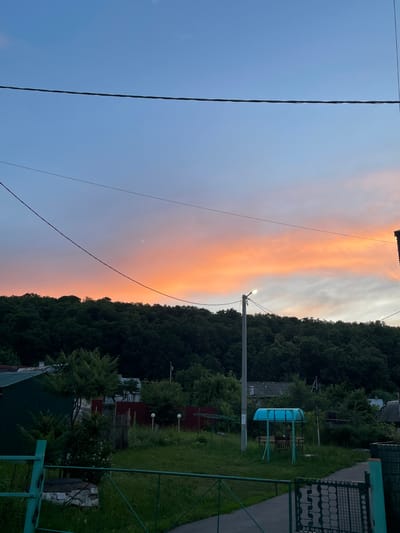 This is a medium-shot, eye-level, outdoor photograph taken in Solnechnaya Polyana, Russia, during a sunset. The sky features a gradient of colors, from a clear blue at the top to vibrant orange and pink hues near the horizon, with scattered clouds. A dark green, dense treeline forms the backdrop, rising up a hill. In the foreground, a dark metal fence runs horizontally across the bottom of the frame. Beyond the fence, a grassy yard is visible with various elements. To the left, a dark green corrugated metal structure and a lighter green metal fence are present. A street lamp stands vertically near the center of the image, with a small, bright light illuminated at its top. To the right of the lamp, a small, bright blue canopy covers a picnic table with benches. Several houses and other structures are partially visible behind the trees and yard. Power lines are strung across the upper portion of the sky. No people are visible in the frame.