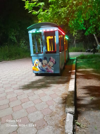 A blue kiddie train ride vehicle with cartoon characters Pluto, Mickey Mouse, and Minnie Mouse painted on its side is parked on a paved walkway at night. The vehicle has colorful LED lights along its roof. The walkway is made of interlocking hexagonal pavers in shades of pink and gray. Green trees with bright green foliage are visible in the background. The scene appears to be in a park or recreational area. A concrete curb runs alongside the walkway. The timestamp "23 июня 2026 г. 22:43" and "Galaxy A12" are overlaid in the bottom left corner of the image.