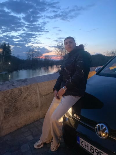 The image captures a young individual, possibly male, with a short haircut, seated or leaning on the front left fender of a black Volkswagen Golf GTD. The individual wears a black puffer jacket with a white marble-like pattern, a black turtleneck, light-colored wide-leg trousers, and white and beige athletic shoes. A silver-toned watch is visible on their left wrist. The car features a partially visible Bulgarian license plate reading "BG KH 895[X]T". The scene is set at twilight in Nevestino, Bulgaria. A stone bridge parapet is visible in the lower left foreground. A calm river extends from the mid-ground towards the horizon, reflecting the sky. The sky transitions from deep blue with stratocumulus clouds overhead to orange and pink hues near the distant horizon, indicative of sunset. Bare deciduous trees and some evergreen foliage line the riverbanks, with mountains visible in the far distance. A distant bridge or structure spans the river, and a small wooden dock is visible on the left bank.