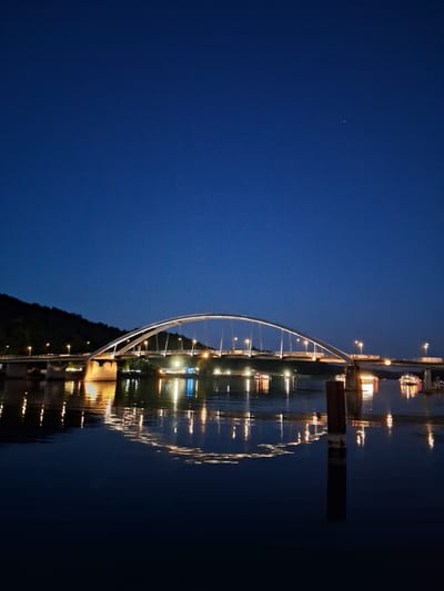 The image shows a wide, arched bridge spanning a dark body of water at twilight. The bridge is illuminated by numerous lights along its length, and its reflection is visible on the calm water's surface. A dimly lit hillside with trees is visible behind the bridge. On the water, several boats with lights are present. The sky above is a deep, clear blue, with a single faint star visible. The scene is located in Vilshofen an der Donau, Germany, and specifically depicts the Vilshofen Bridge over the Danube River.