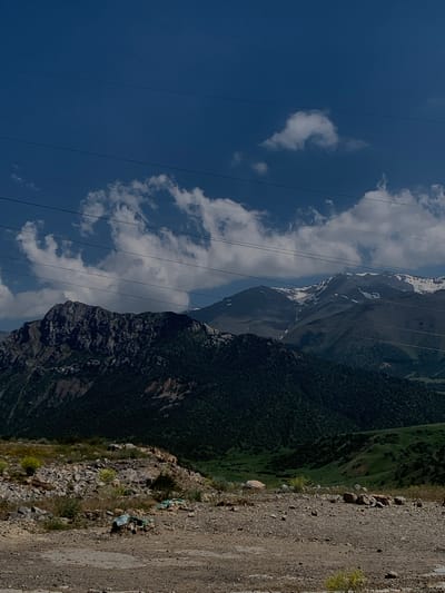 The image depicts a mountainous landscape under a clear blue sky with scattered white clouds. Power lines are visible in the upper portion of the frame, stretching horizontally across the sky. In the mid-ground, dark, densely vegetated mountains rise, with a rocky, less vegetated peak in the left foreground. The background reveals taller, snow-capped mountains. The foreground consists of a dirt and gravel ground with scattered rocks and dry vegetation. There are no people or specific actions depicted. The scene appears to be a natural environment, likely in the vicinity of Dardar, Uzbekistan, given the mountain ranges and vegetation typical of the region.