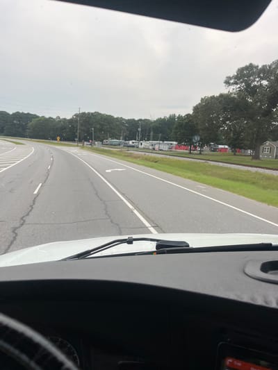 The image shows a view from inside a vehicle looking out through the windshield. The vehicle is on a road with multiple lanes, indicated by white lane markings. The road curves to the left in the foreground. On the right side of the road, there is a grassy area with train tracks running parallel to it. Beyond the tracks, there are several buildings, some of which appear to be storage units or small businesses with red siding. Further in the distance, there are trees and a cloudy sky. The vehicle's dashboard is partially visible at the bottom of the image, with a steering wheel on the left. There are no people visible in the image. The scene suggests a rural or semi-rural area.