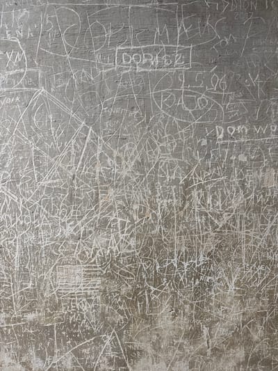 A gray concrete wall covered with white scratch marks. The marks are names and dates, including "Tom 2015," "2017," and "Kevin." The coordinates are 50.033 latitude, 19.179 longitude. Brzezinka, Poland. The wall is located in the former Auschwitz II-Birkenau concentration camp in Oświęcim, Poland, also known as Brzezinka, where a former German prisoner of war left his mark.
