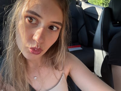 The media file is a close-up, rotated selfie of a young woman. She is in a car, suggested by the black leather car seats visible in the background. The woman has long, light brown hair that is falling around her face. She is wearing a light-colored top and a delicate necklace with a small, sparkling pendant. Her lips are slightly parted, and she has on light pink lipstick. The lighting suggests it is daytime, with sunlight casting shadows on her face and illuminating her hair. The woman's expression is somewhat playful or alluring. No text is visible in the image. The location context is Iffezheim, Germany.