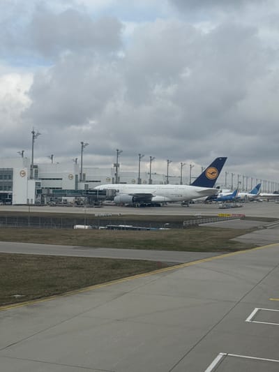 The image presents an exterior view of Munich Airport in Oberding, Germany, on an overcast day. A prominent white Lufthansa Airbus A380 aircraft is parked at a gate in the mid-ground, connected to the terminal by a jet bridge labeled "Revolut." Ground support equipment surrounds the A380's forward fuselage and underwing areas. The multi-story white terminal building, featuring the Lufthansa crane logo, occupies the left and background, equipped with multiple jet bridges and tall light poles. Other aircraft, including one with a blue tail and another with a blue and green tail design, are visible parked further down the tarmac. The foreground and mid-ground consist of concrete tarmac with yellow and white markings, bordering sections of sparse, dry grass. The sky is uniformly gray with extensive cloud cover.