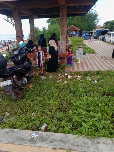 The image shows a group of people gathered under a wooden shelter at a beachside location in Pameungpeuk, Indonesia. There are six people in the image, including a man wearing a blue shirt and a red hat, a woman wearing a black hijab, a young boy wearing a blue and white striped shirt, a young girl wearing a pink and black dress, and two women wearing headscarves. The man is sitting on a motorcycle, which is parked next to the shelter. The other individuals are standing or sitting at a table underneath the shelter. There is a lot of litter on the ground in front of the shelter, mostly consisting of plastic bottles and bags. The environment is a grassy beachside area with palm trees in the background. The ground is a mixture of grass, sand, and concrete. The shelter is made of wood and has a concrete roof. There is a blue plastic container next to the table, and the individuals appear to be engaged in a conversation or interaction. The shelter is located on a concrete path that leads to the beach. The image was taken during the day, as the sun is shining and there are no shadows. The image is a candid shot, as it appears to have been taken without the subjects being aware of the photographer.
