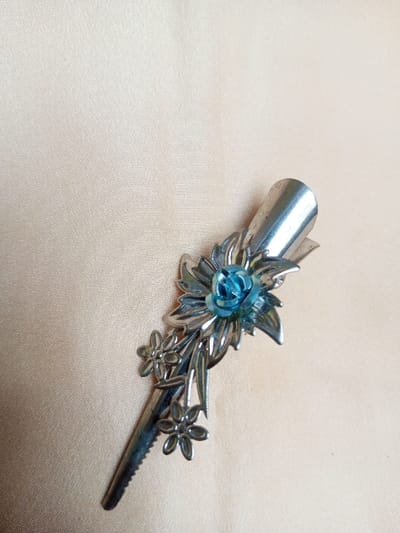 A single metallic hair clip is depicted at an oblique angle, positioned from the lower-left to the upper-right of the frame. The item is silver-toned, approximately 8-10 cm in length, and features an embossed floral design. A central element consists of a small, molded, turquoise-blue rose, surrounded by larger silver-colored leaves and petals. Below this main motif, a cluster of smaller, simpler silver-colored flowers and leaves extends along the clip's body. The bottom section of the clip exhibits a serrated edge, consistent with a hair-gripping mechanism. The object rests on a flat, light beige textile surface with a subtle weave texture. The scene lacks any human presence or discernible action, and is situated within Qarabucaq qəsəbə, Azerbaijan.