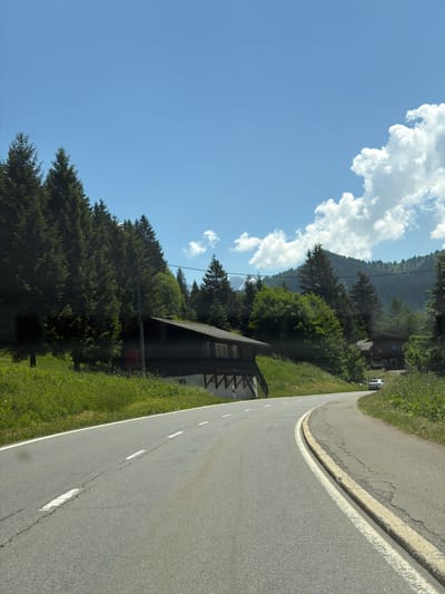 This media file captures a scenic view of a winding road in Morgins, Switzerland. The road, with white lane markings and a yellow dividing line, curves through a lush green landscape. On the left side of the road, a dark-colored building with a pitched roof and a single window is nestled amongst dense evergreen trees that climb up a steep mountainside. The trees create a rich, dark green backdrop against a bright blue sky dotted with fluffy white clouds. The sun appears to be high in the sky, suggesting it is daytime, and the weather seems fair and clear. The scene evokes a sense of peacefulness and natural beauty, characteristic of a mountainous region. There are no people or animals visible in the frame, and no discernible text. The perspective suggests the photo was taken from a moving vehicle, possibly a car or bicycle, given the close proximity to the road and the slight tilt of the image.