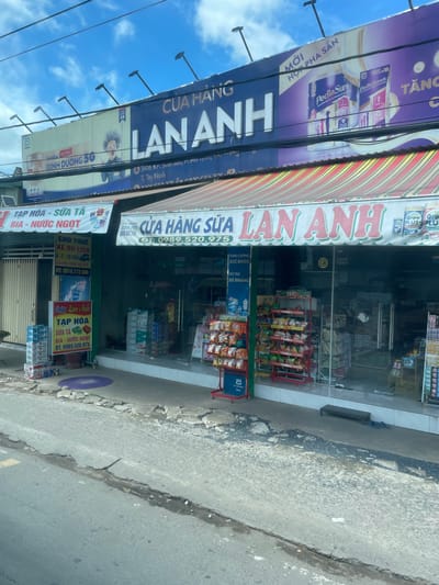 The image shows a street view in Phường An Tịnh, Vietnam, featuring a storefront labeled "CỬA HÀNG SỮA LAN ANH". The store is a convenience store or a milk shop, indicated by the display of various products like bottled drinks, snacks, and milk powder cans visible through the glass doors and on shelves outside. Above the store, a large billboard advertising "DINH DƯỠNG 5G" and "Pediasure" is present. The street in front of the store is paved with asphalt, showing signs of wear and tear with cracks and uneven sections. There are some parked vehicles and stacks of beverages, likely for sale, outside the store. The overall scene depicts a typical commercial area in Vietnam.