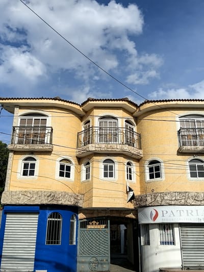 This is a medium-angle, eye-level, exterior shot of a three-story building in Puerto Plata, Dominican Republic. The building is painted a bright yellow and has a textured stucco finish. The roof is tiled in a reddish-brown color.
The building has multiple windows, many of which are arched and have white frames and dark panes. Some of the windows on the upper floors open onto small balconies with ornate black metal railings. The ground floor features a vibrant blue facade on the left, with a corrugated metal roll-up door and two arched windows with metal bars. To the right of the blue section, there is an arched entranceway with a green metal gate. Further to the right, a white commercial unit with a sign for "PATRIA SEGUROSPATRIA.COM" is visible. The sign has a logo with red and white curved elements.
The sky above is a bright blue with scattered white clouds. A black electrical wire runs diagonally across the upper portion of the image. A small black and white flag or banner hangs from the yellow facade of the building near the center. The overall scene depicts a commercial and residential building in what appears to be an urban setting. No people are visible in the image.
