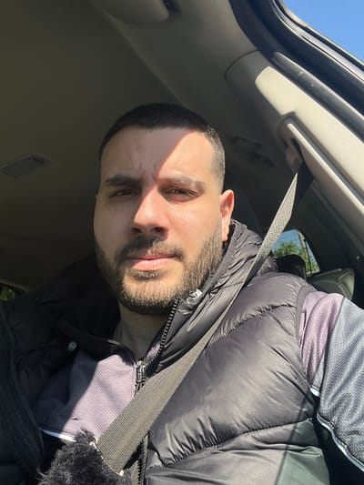 A medium shot depicts a man with short dark hair and a beard, seated in a car. He is wearing a black puffer jacket and a seatbelt. The car's interior is visible, including the ceiling and dashboard. Sunlight is streaming into the car, casting shadows on his face. The location of the photo cannot be verified as Trastikovo, Bulgaria, from this image.