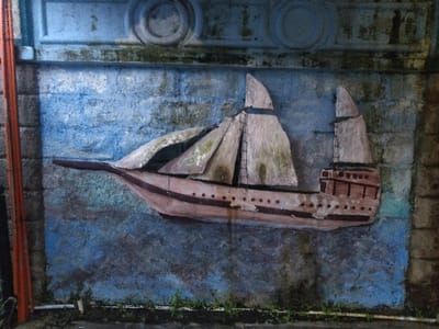 The image displays a weathered, three-dimensional relief sculpture of a sailing ship affixed to a painted concrete wall. The specific location within Ciudad de San José, Costa Rica, cannot be determined from the image alone.
The ship, depicted in profile and oriented towards the left, features a pointed bow, a prominent hull painted in dark reddish-brown with lighter horizontal bands and rows of circular indentations, and a raised multi-tiered sterncastle. Two masts are visible, each supporting a large, partially furled sail rendered in a light, discolored material, exhibiting green and grey biological growth.
The wall behind the ship is painted with a maritime scene, presenting a dark blue and grey lower section suggestive of water, transitioning to a lighter blue upper section. The entire wall surface is rough-textured and shows significant wear, with areas of faded paint, chipping, and widespread dark grey and green biological growth. A horizontal architectural frieze with recessed circular motifs runs along the top edge of the wall.
To the far left of the frame, a vertical reddish-orange pipe or conduit is visible, partially obscured, running parallel to a darker vertical structure. At the base of the wall, a narrow strip of dark ground is present, from which small patches of green vegetation protrude. The scene appears to be outdoors, under muted lighting conditions. No human subjects are present.