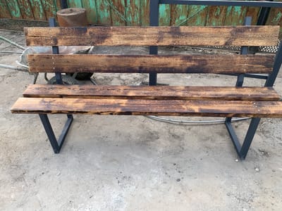 A rustic wooden bench with three slatted seats is placed on a dirt and gravel surface in front of a weathered green metal fence. The bench has black metal V-shaped legs. In the background, behind the bench, is a metal wheelbarrow and a large cylindrical object. The scene appears to be an outdoor area in a city, possibly a park or industrial yard in Berëzovaya Roscha, Russia.