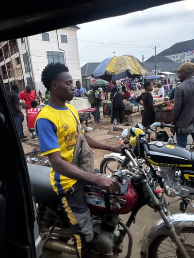 The image depicts a bustling street market scene in Obinze, Nigeria. In the foreground, a young man wearing a yellow and blue t-shirt and dark pants is seated on a red and black motorcycle. He has a backpack slung over his shoulder and appears to be looking to his right. Another motorcycle, primarily black with yellow accents and the brand name "DAYLONG" visible, is parked alongside his.
The background reveals a crowded market with numerous people engaged in various activities. Stalls with colorful umbrellas are set up, suggesting the sale of goods. Buildings, including a multi-story structure with balconies under construction and a white building with multiple windows, are visible in the distance. The ground is unpaved and appears dusty, with some debris scattered around. The sky is overcast, indicating a cloudy day. The overall impression is one of a vibrant and active community market.
