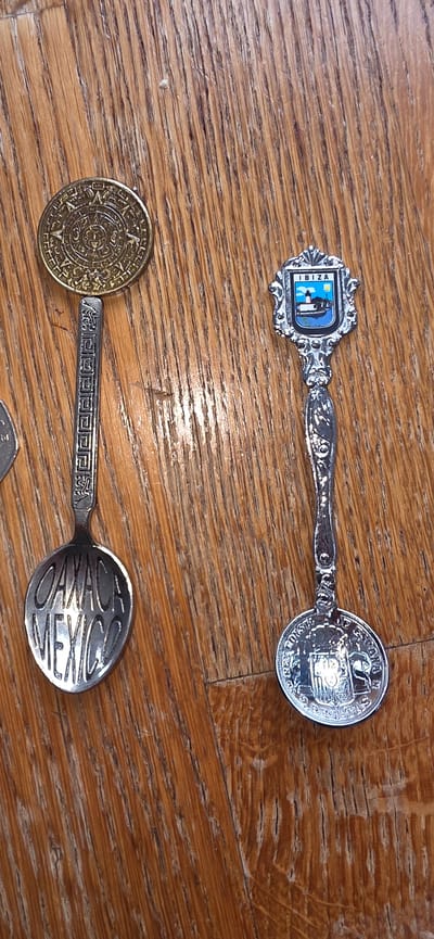 Two souvenir spoons are displayed on a wooden surface. The spoon on the left has a coin with a sun symbol at its head, and the spoon's bowl is inscribed with "OAXACA MEXICO." The spoon on the right has a shield emblem featuring a lighthouse and a body of water, with "IBIZA" written above it. The wooden surface appears to be a table or floor with prominent grain patterns. The image is an overhead shot. There are no people visible in the image, and no indication of the city of Marum, Netherlands.