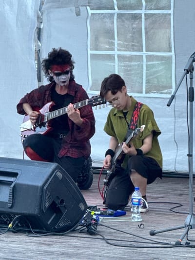 Two individuals are performing music on a wooden stage. The individual on the left, with black curly hair and face paint consisting of white, black, and red, is wearing a dark plaid shirt over a black t-shirt and black pants. They are kneeling and playing a red electric guitar. The individual on the right, with short dark hair and glasses, is wearing a green short-sleeved shirt with a red and black flame pattern strap, black shorts, and black wristbands. They are also kneeling and playing a black electric guitar. A large black speaker is positioned in the foreground to the left, and various cables and a bottle of water are visible on the stage. Behind the performers is a white tent-like structure with grid-like window patterns. The scene appears to be outdoors, likely at an event or festival. There is no indication that this scene is located in Votkinsk, Russia.