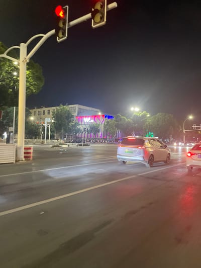 The image depicts a nocturnal street scene in Dali City, China, facing approximately east towards Dali Wanda Plaza. The multi-lane asphalt road, which appears wet, occupies the foreground and midground. Overhead, a traffic light pole with two illuminated red signals indicates a stop.

To the left, set back from the road, is the prominent **Dali Wanda Plaza** building, identifiable by its large, illuminated "W" logo displayed in red, white, and blue colors. Adjacent to the plaza on the far left is a multi-story building with multiple windows. A white barrier with red warning stripes stands on the extreme left near the sidewalk. Several motorcycles or scooters are parked in front of the Wanda Plaza building.

In the midground, a light-colored SUV, likely white or silver, with a green new energy vehicle license plate, is visible from the rear, its taillights illuminated red. It is positioned in one of the road lanes, facing away from the camera. To its right, a portion of a red vehicle's rear, also facing away with illuminated red taillights, is visible on the far right edge of the frame. Streetlights illuminate the scene, casting reflections on the wet road surface. No discernible people or specific interactions are visible.