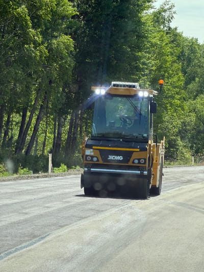 The image shows a yellow XCMG soil compactor operating on a paved road under construction, likely in the city of Aur, Russia. The road surface is a mixture of asphalt and gravel, indicating ongoing road work. Lush green trees line the right side of the road, with a clear sky above. The compactor's cabin is visible, with a driver inside operating the machinery. Bright LED lights on the compactor are illuminated, and an orange flashing beacon is present on top. The front drum of the compactor is in contact with the road surface, performing its compaction duties. The overall scene depicts a typical construction site with heavy machinery in operation.