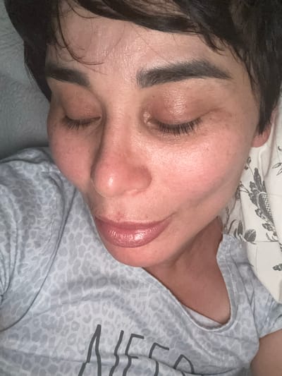A close-up, overhead shot shows a woman with her eyes closed, lying on her side on a bed with a patterned pillow. She has short, dark hair, dark eyebrows, and appears to be wearing light-colored makeup, including lipstick. Her skin has a slightly flushed appearance and some texture. She is wearing a light gray t-shirt with a subtle animal print and appears to be indoors. The background consists of a textured gray surface and the floral patterned pillow. There is no indication of the city of Bellmore, United States of America.