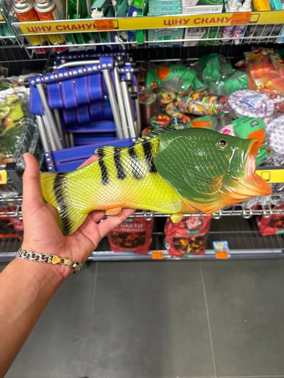 A person's hand, wearing a silver chain bracelet, holds a brightly colored toy fish with an open mouth. The toy is green on its head and orange on its mouth, with a yellow body and black stripes. It is positioned in front of a retail display shelf filled with various products. In the background, there are blue chairs stacked, and shelves with packaged goods, some with yellow signage that reads "ЦІНУ СКАНУЙ". The scene appears to be in a store.