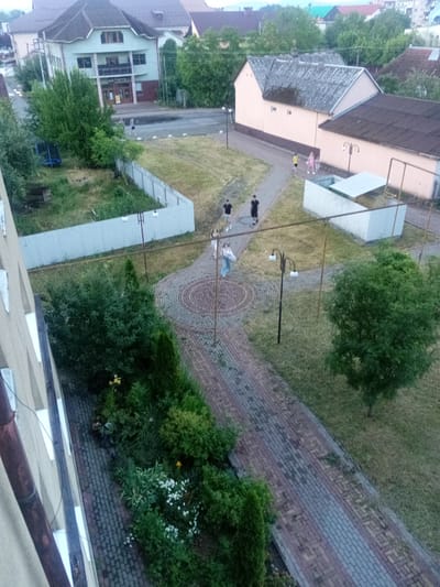 The image depicts a courtyard area in Tiachiv, Ukraine. In the background, a two-story building with a light green facade and a dark roof is visible. Adjacent to it, a lighter colored building with a grey roof and a pinkish facade runs parallel to the foreground. The courtyard itself is characterized by a paved walkway made of red and grey bricks, bordered by green grass and trees. Several people are present: two individuals in dark t-shirts and shorts walk on the paved path in the upper middle of the image. Further down the path, another individual in a light-colored shirt and jeans walks away from the camera. To the right, three younger individuals are seen walking together, two appearing to be children or teenagers dressed in light-colored clothing. A blue trampoline is situated in the grassy area to the left, beyond a light-colored metal fence. The overall scene suggests a residential or communal outdoor space.
