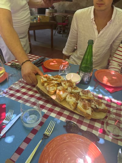 In an outdoor dining setting in Agra, Switzerland, a table is laden with food and drinks. A person's hand, adorned with a beaded bracelet, reaches towards a wooden board showcasing numerous slices of bread topped with white cheese and fried onions. Nearby, two red plates, a green bottle of Valser water, and several glasses are visible. The table is covered with a red and white checkered tablecloth and a blue placemat. In the background, other diners are partially visible, along with wooden furniture and barrels, suggesting a rustic ambiance. The lighting indicates it is daytime, with sunlight casting shadows on the table.