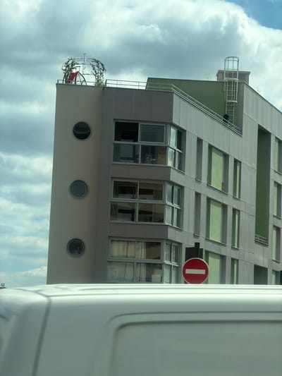 The media file shows a modern building with a gray facade and green-tinted windows. The building has several floors, with a balcony on the lower level featuring a decorative, crown-like structure adorned with vines and a red chair. Three circular windows are visible on the lower section of the building. A red and white "no entry" sign is mounted on the building's facade. The sky in the background is cloudy. The image appears to have been taken during the daytime. A portion of a white vehicle is visible in the foreground, partially obscuring the view of the building. The image is oriented vertically, with the building appearing to lean to the left. Based on the provided location context, this scene is in Alfortville, France.