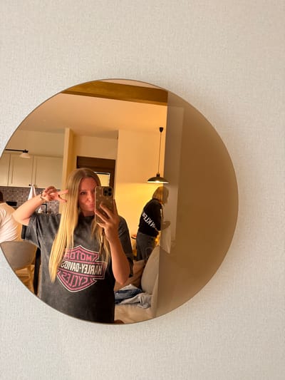 A woman with blonde hair, wearing a dark t-shirt with a pink Harley-Davidson logo, takes a mirror selfie. She is holding her phone in her right hand and making a peace sign with her left hand. In the reflection of the round mirror, another person wearing a black sweatshirt is visible in the background, looking down. The setting appears to be an indoor space, possibly a room in Kovačka Dolina, Montenegro, with furniture and a lamp visible in the reflection. The wall is textured and light-colored.