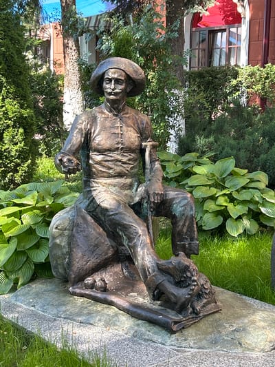 This is a photograph of a bronze statue of a man sitting on a rock, in a garden setting. The man is wearing a hat, a buttoned shirt, and trousers. He is leaning on a cane with his left arm and holding something in his right hand. The statue is positioned on a stone pedestal, surrounded by lush green plants and grass. In the background, there is a building with a red facade and windows, and trees. The photograph is taken in the city of Oktyabrskoye, Russia.