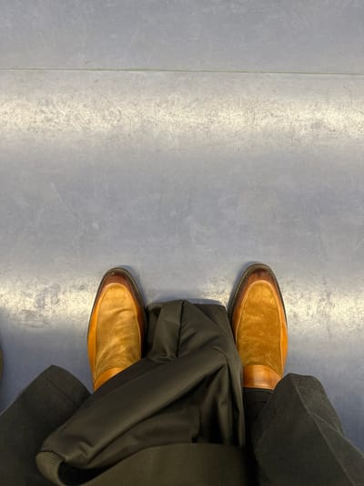 A person is wearing a black jacket and black pants, with both feet on the ground, wearing brown leather shoes. The scene is inside, on a floor that appears to be concrete. This is a shot from above the person, from their own perspective. The image was likely taken in the town of Mandello del Lario, Italy, based on the given coordinates. The lighting in the image is dim and slightly yellow. The person is not interacting with anything.