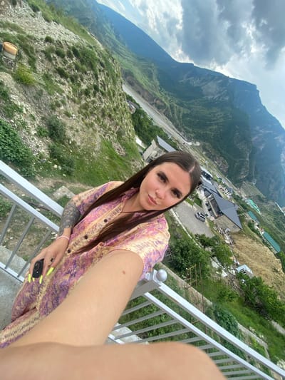The image shows a woman taking a selfie from an elevated position overlooking a village nestled in a mountainous valley. The woman is wearing a patterned dress and has long brown hair. She is holding a black object in her left hand, which appears to be a camera or phone. Her nails are painted neon yellow. The background displays a steep, green mountainside with scattered buildings and a winding road. The sky is overcast with dramatic clouds. The location appears to be the village of Chalda, Russia, known for its mountainous terrain.