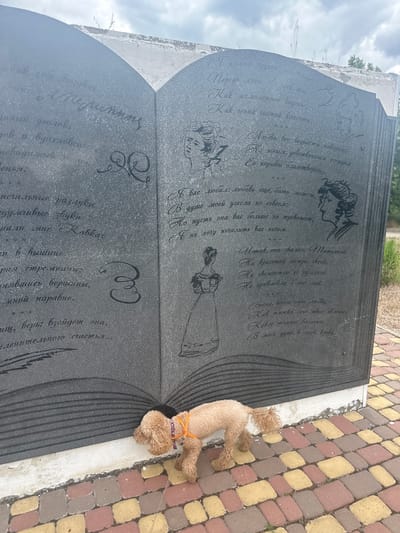 The image shows a gravestone with carved text and portraits. A small, light brown poodle is lying on the ground next to the gravestone, facing away from the camera. The gravestone is made of dark stone and is etched with what appears to be poetry and decorative elements, including profiles of people and floral designs. The text is in Cyrillic script. The setting appears to be an outdoor cemetery, with a cobblestone path made of red, yellow, and gray stones visible to the right of the gravestone. In the background, there are hints of greenery and a cloudy sky, suggesting it might be daytime with overcast weather. The overall mood is somber, as is typical of a memorial site, with the presence of the dog adding a touch of unexpected companionship or a sense of a visit. The text on the gravestone is too small and blurry to be fully deciphered in the main image, but the provided crops offer a closer look at some lines of poetry, suggesting themes of love, memory, and perhaps loss. The location context provided indicates Novokubansk, Russia.