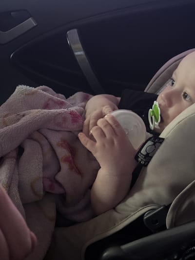 A close-up, dimly lit image shows a baby or toddler seated in a car seat, likely inside a vehicle, in Klenovskoe, Russia. The child, visible from the chest up, has light skin and is wearing a black top. A green and white pacifier is in the child's mouth, and their hands are clasped around a white baby bottle or sippy cup. The child's eyes are open, looking towards the upper left of the frame.
The baby is partially covered by a soft, plush blanket, which is pinkish with subtle patterns in shades of pink, red, and yellow. The car seat itself is beige or light brown, and a label on its side is visible, displaying text that includes "S.H.P" and "SIDE IMPACT PROTECT", indicating a safety feature. The background reveals parts of the car's dark interior, possibly a door panel or console, with some silver-colored trim elements. The lighting suggests an indoor setting or a low-light outdoor condition, creating a somewhat cozy atmosphere around the child. No specific activity beyond holding the bottle and being wrapped in the blanket is distinctly identifiable; the child appears calm and settled.