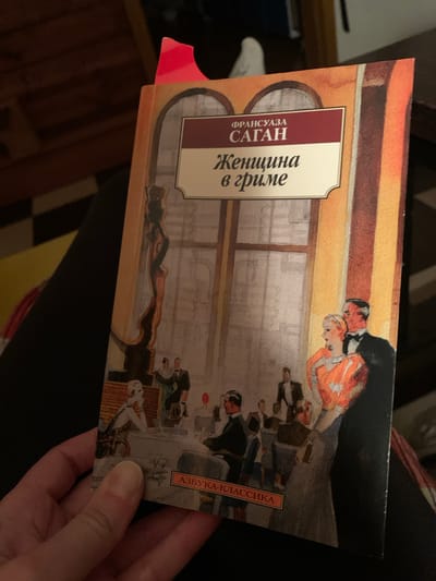 A close-up shot shows a book cover held in a person's hand. The book is titled "Женщина в гриме" (Woman in Makeup) by Франсуаза Саган (Françoise Sagan). The book belongs to the "Азбука-классика" (Azbuka-Klassika) series. The cover illustration depicts a scene with several figures in what appears to be a social gathering, possibly in a 1920s setting. A couple is in the upper right, and seated people are positioned below near a low table. The background shows a room setting with window detailing. The book is slightly tilted. Kremyonki, Russia, is not mentioned in the text on the book cover nor depicted in the cover's scene.