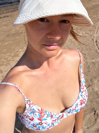 A woman wearing a white bucket hat and a floral bikini is standing on a sandy beach, with the water visible in the background. The image is a close-up, focusing on her upper body and face. The setting appears to be a sunny day. The beach is part of the coastline of Kalngale, Latvia.