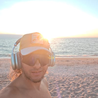A close-up, eye-level, outdoor selfie shows a person wearing headphones and a yellow baseball cap with a white brim and a patch on the front. The person is also wearing large, dark sunglasses. The background consists of a calm ocean with gentle waves and a sandy beach. The sun is setting or rising, casting a warm glow over the scene and creating a bright light that is partially obscuring the sky. The sand on the beach is light-colored and appears to have small shells or pebbles mixed in. The person's shoulders and part of their chest are visible, and they have a beard. The overall mood is relaxed and peaceful, suggesting a moment of personal enjoyment by the sea. The location is stated as Chelyadinovskoye Rural Settlement, Ukraine, which is unlikely given the presence of a large body of saltwater and beach.