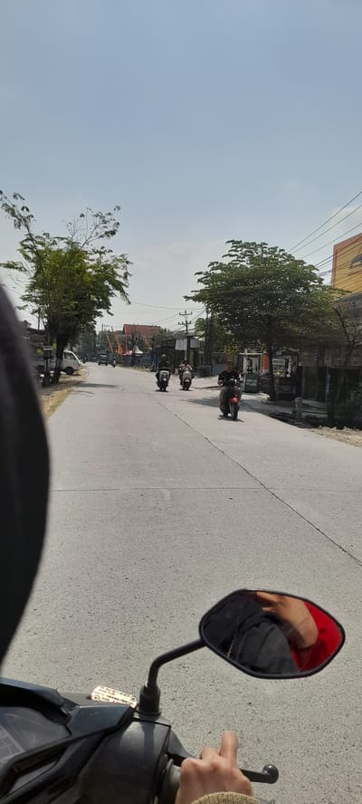 The image captures a view from the perspective of someone riding a motorcycle on a paved road, likely in Grabagan, Indonesia, given the context. The sky is a clear, pale blue with a few faint clouds. On the left side of the road, there's a white pickup truck parked, and behind it, a building with a sign that reads "PARKING." A large tree with sparse foliage stands near the road.

Further down the road, several other motorcycles are visible. One is ridden by a person wearing a dark helmet and a black shirt. Another motorcycle, a white scooter, is being ridden by someone in a dark outfit. Further ahead, a person on a red motorcycle is visible, wearing a black t-shirt and shorts.

On the right side of the road, there are shops and buildings. One building has a bright yellow facade and an orange awning. Further down, there is a gas station with green pumps. Another building has a sign that appears to be in Indonesian, but the text is too small to read clearly. The overall environment suggests a rural or semi-urban area with some commercial activity.

The reflection in the motorcycle's rearview mirror shows the arm and shoulder of the rider wearing a red garment and a black jacket, along with a bracelet. The motorcycle's handlebar and dashboard are also visible in the foreground. The road has a visible line dividing the lanes and appears to be in good condition. The presence of flags or banners, possibly for a local event or celebration, is noted on the left side of the road in the distance.
