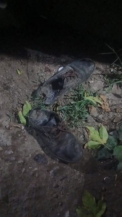 The image shows a pair of worn, dark-colored shoes with laces, lying on the ground amidst sparse vegetation and dirt. The setting appears to be outdoors. There are no people present. The location is identified as Putucual, Venezuela, but there are no specific landmarks or indicators of the city visible in the image.