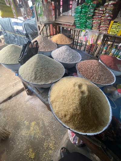 The image depicts a market stall in Ojodu, Nigeria, displaying various grains and beans. Multiple metal bowls contain rice, beans, and other legumes of different colors and textures. Packaged goods are visible on shelves in the background. A broom lies on the floor. A foot wearing a sandal is visible in the corner. The floor has scattered grains and debris.