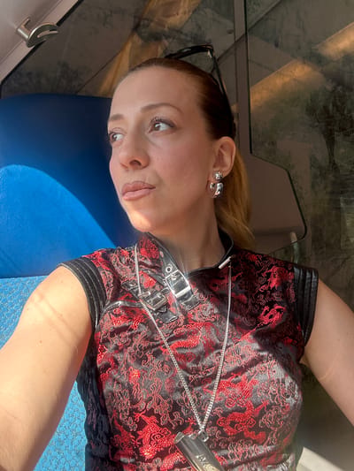 In this medium shot, a woman lies on her side, her body angled towards the viewer. She is wearing a black and red patterned garment adorned with intricate dragon designs. The fabric has a slight sheen, suggesting it might be silk or satin, and features a prominent zipper and buckle details at the neckline. A silver chain necklace rests on the garment. The woman's head is tilted back, and her eyes are wide open, looking slightly to her right. Her lips are parted, and her expression appears surprised or intrigued. She has on silver, flower-shaped stud earrings. Her blonde hair is pulled back. A pair of black-rimmed glasses are pushed up on her forehead, resting near her hairline.
The background is blurred, but hints of a reflective surface, possibly glass, are visible at the top. Sunlight casts a sharp shadow across the woman's chest and arm, indicating a bright, sunny day. She is lying on a textured blue surface, which could be a cushion or upholstery. The overall impression is that of a candid moment captured, possibly during leisure or travel. There is no visible text in the image. The setting is the Wassenaar, Netherlands.
