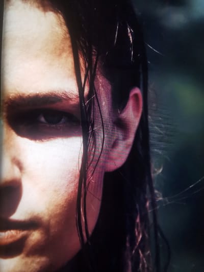 The media file is a close-up, side-profile shot of a person's face, with only the left side of their face visible. The person has long, dark, wet hair that is partially obscuring their face. Their left eye is visible, with dark eyebrows and eyelashes. The left ear is also prominent in the frame. The skin tone is fair, with warm lighting highlighting the contours of the face. The background is dark and out of focus, suggesting an outdoor setting, possibly at dusk or in shaded conditions. There are no visible objects or text in the image. The overall mood conveyed is somewhat somber and introspective, possibly due to the lighting and the person's direct gaze. The location context provided is Serov, Russia, but no specific cues within the image directly confirm this.