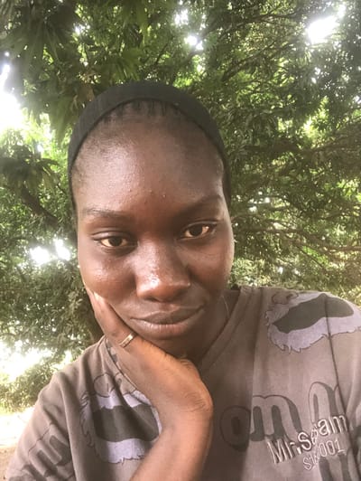 A headshot-style image depicts an adult female outdoors in Ninzo South, Nigeria. The subject has a dark complexion, is wearing a black headband, and a dark grey t-shirt with light grey patterns and partially visible text "Mr. Spam S/W 001." A plain gold-colored band ring is present on her left ring finger. Her left hand is raised, with fingers visible, supporting her chin. She maintains direct eye contact with the camera. The background consists entirely of dense green tree foliage, with bright sunlight filtering through the leaves, indicating an outdoor setting under a canopy.