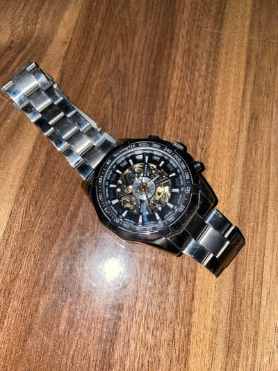 A silver watch lies on a wooden surface. The watch has a black dial with intricate mechanical details visible through the face. Markings such as "TACHYMETER," "WINNER," "WATCH," and "SPORT" are printed on the dial. The band appears to be made of silver metal links. The watch's position on the wood is slightly diagonal, with a bright spot reflected on the surface behind it.
