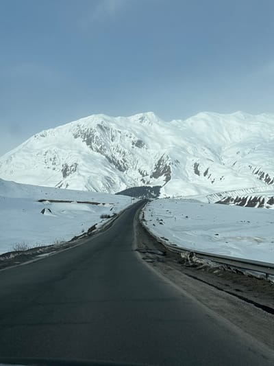 The image depicts a straight, two-lane asphalt road leading into the distance, ascending slightly towards a mountainous landscape, specifically located in Sioni, Georgia. The road surface appears generally clear, with some minor dark patches and irregularities visible along its right edge where it meets a metal guardrail. Both sides of the road are covered in significant amounts of snow, forming gentle slopes and drifts that extend into the midground.

In the midground, the snow-covered terrain features undulating hills, with patches of darker, exposed earth or rock visible in some areas. A small cluster of dark evergreen trees is discernible on the left side, further down the road's path, and a single utility pole stands vertically near the road, also on the left.

The background is dominated by an expansive range of high, rugged mountains completely blanketed in white snow. The mountain slopes show variations in texture, with some areas appearing smooth and others exhibiting more defined ridges and valleys, suggesting rock formations beneath the snow. The sky above the mountains is a clear, pale blue, with no discernible clouds. There are no individuals, vehicles, or animals visible in the scene, and no overt actions or interactions are depicted. The overall environment suggests a cold, winter setting.