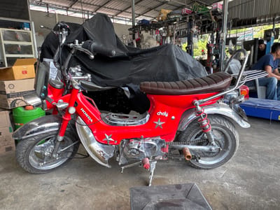 The image shows a red Honda Chaly mini bike parked in what appears to be a workshop or garage. The motorcycle is partially covered with a black tarp. In the background, there are shelves filled with various items, tools hanging on the wall, and two men are seated, one of whom is looking towards the motorcycle. The setting suggests a commercial or repair environment, likely in ตำบลเกาะพะงัน, Thailand, given the context provided. The floor is concrete, and the overall lighting indicates an indoor space with some natural light filtering in.