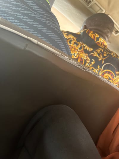 This image captures the interior of a vehicle in Shuwarin, Nigeria, from the perspective of a seated passenger. In the lower left quadrant, a knee covered in dark, likely black, fabric is visible. Adjacent to it on the right, a folded piece of orange-brown fabric is present. The midground features the back of a dark, possibly black or dark gray, upholstered seat, which occupies a significant portion of the frame horizontally. The top edge of this seat displays evident wear, with white or light-colored material exposed along the seam. Beyond this seat, in the upper right background, the back of an individual's head and upper torso are visible. This person is wearing a black shirt patterned with an ornate gold and orange floral and scroll design. To the upper left, a portion of another seat's back, featuring a gray and black geometric or herringbone pattern, is visible. A thin strip of bright green material is noticeable near the top edge of this patterned seat. The vehicle's light-colored ceiling, possibly beige or off-white, with a recessed light or vent panel, is visible in the upper background.