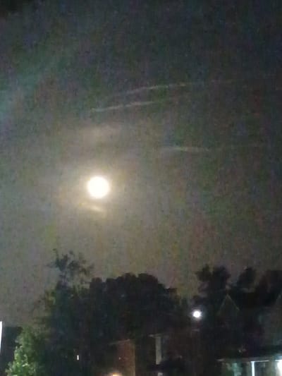 A night sky scene is depicted, with a bright, glowing orb, presumably the moon, positioned in the upper left quadrant. The moon is surrounded by a hazy, slightly luminous atmosphere. Streaks of light, possibly from aircraft contrails or clouds, are visible in the sky above the moon. The lower portion of the image is dominated by silhouettes of trees and the outlines of buildings, suggesting an urban or suburban environment. The overall lighting is dim, characteristic of nighttime. There is no discernible activity of people or specific objects that would pinpoint a precise location within Nicholasville, United States of America, beyond the general context of a sky view over a populated area.
