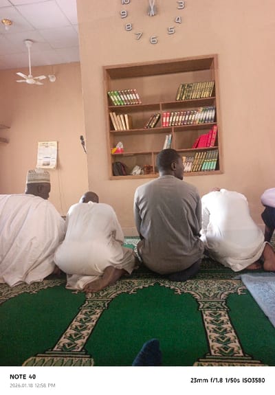 The image depicts an indoor scene within a mosque or prayer room in Tudun Wazurchi, Nigeria. Four adult males are observed from behind, kneeling or sitting on their heels on green prayer rugs. Three individuals on the left are dressed in white traditional robes; the leftmost man wears a white skullcap, and the second from the left is bald. The third man from the left, positioned centrally, wears a grey long-sleeved shirt and dark trousers. The fourth man on the far right also wears a white robe and is bowing his head. A fifth individual's foot, wearing a blue sock, is visible in the bottom right foreground, indicating the photographer's kneeling position.

The room features light brown walls and a white grid-patterned ceiling. A white, three-bladed ceiling fan and a circular golden light fixture are mounted on the ceiling to the left. A large, frameless wall clock with silver numerals and hands, indicating approximately 12:58, is affixed to the upper right wall. Below the clock, a wooden bookshelf with multiple compartments is mounted, containing numerous books, predominantly Islamic texts with covers in green, gold, red, blue, and white. A white tissue box and a dark bag are also on the shelves. A white document with Arabic script, possibly a calendar or religious poster, is visible on the left wall, above a partially obscured black microphone on a stand. The floor is covered with green prayer rugs featuring intricate patterns of arches, pillars, and geometric designs in white and gold. The overall scene suggests a moment of prayer or religious study. The image metadata indicates it was captured on 2026.01.18 at 12:58 PM.